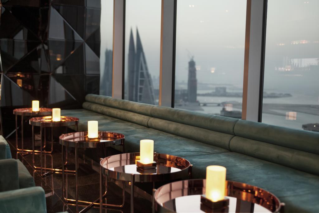 The Domain Bahrain Hotel And Spa - Adults Friendly 16 Years Plus Manama Ngoại thất bức ảnh The photo depicts a stylish lounge area with a modern aesthetic. There are several circular tables made of metallic materials, each topped with a small glowing candle. A long, plush seating area in a teal color runs along the window, offering a view 
